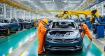 The image shows a bustling automotive factory floor with workers assembling cars on the production line. Numerous vehicles in various stages of assembly are visible, along with heavy machinery and equipment.